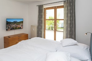 A bedroom with a bed, a wooden dresser, a radiator, a television on the wall, and a window with curtains.