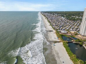 Uma área costeira com praia, estacionamento e um edifício grande.