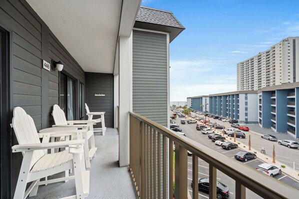 A balcony with white Adirondack chairs, a view of a parking lot, and a high-rise building in the background. - Myrtle Beach Resort Ocean View Condo (Myrtle Beach)