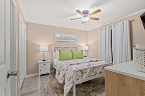 Quarto com cama, mesas de cabeceira, ventoinha no teto, um quadro e uma janela com cortinas.