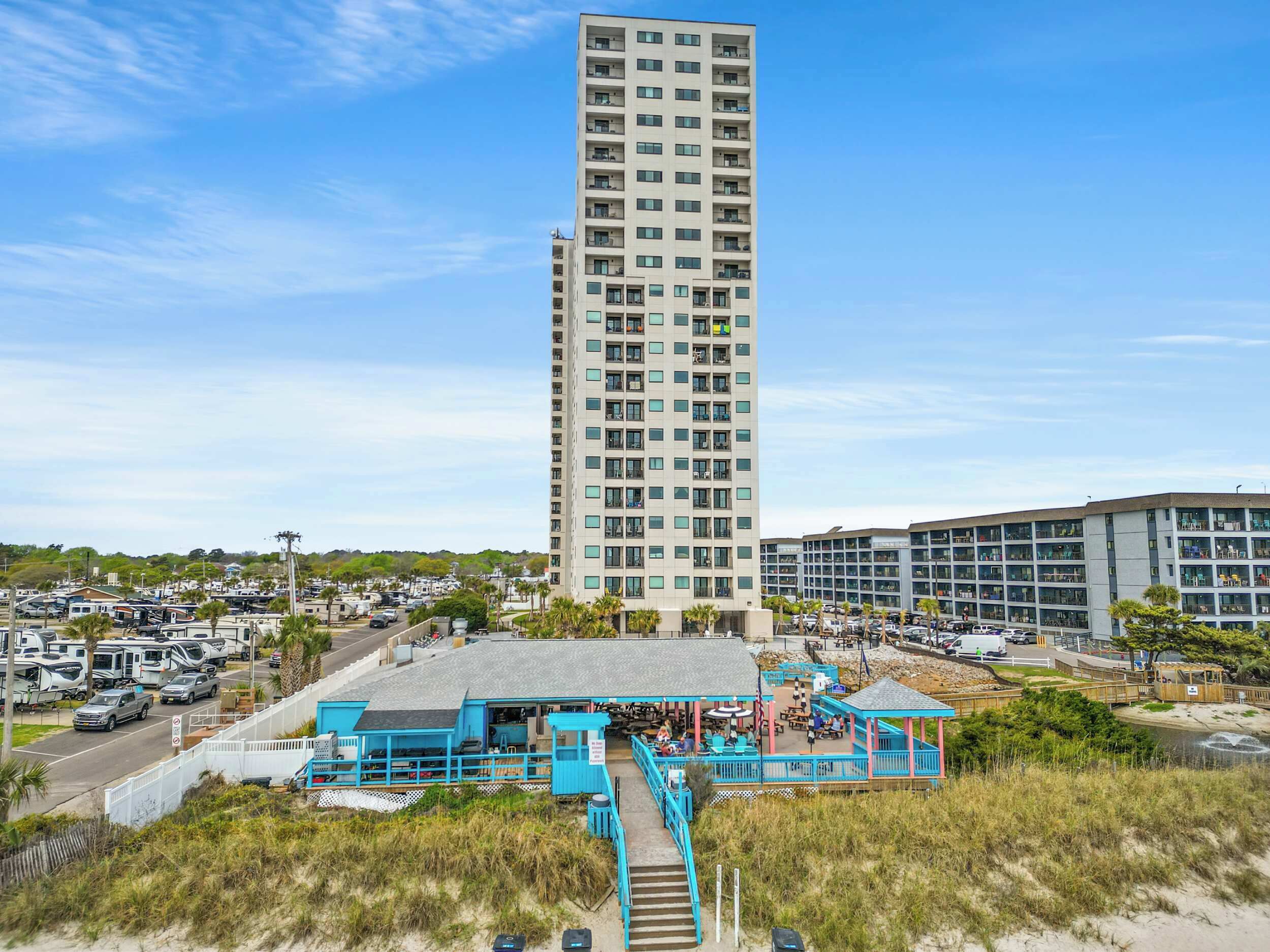 A high-rise building with multiple floors, a beachside restaurant with outdoor seating, and a parking area with RVs.