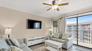 A modern living room with a flat-screen TV, a ceiling fan, a sofa, a coffee table, and a balcony with a view of the buildings. - Myrtle Beach Resort Ocean View Condo (Myrtle Beach)