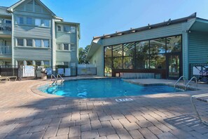 Uma piscina com deck de tijolos, rodeada por um edifício com grandes janelas e área coberta.