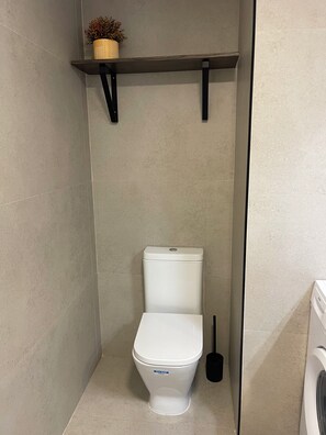 A compact bathroom with a white toilet, a shelf above it, and a washing machine to the right. - STUDIO IN ALICANTE CITY CENTER (Alicante Ciudad)