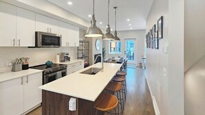 Een moderne keuken met een centraal eiland, roestvrijstalen apparatuur, hanglampen en uitzicht op de eetruimte.