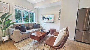 Een moderne woonkamer met een bank, salontafel en een fauteuil. Er is een groot raam, een flatscreen-tv en een koelkast.