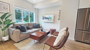 A modern living room with a sofa, coffee table, and a chair. There's a large window, a flat-screen TV, and a refrigerator. - Pet-Friendly Panorama Escape / Hot Tub / Sleeps 10 (Panorama)