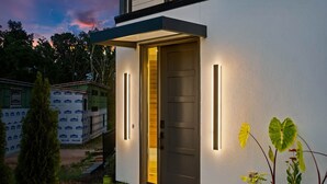 Ein modernes Haus mit einer dunklen Tür, zwei vertikalen Lampen und einem kleinen Garten mit einer Pflanze.
