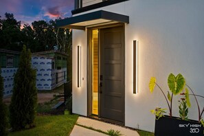 Ein modernes Haus mit einer dunklen Tür, zwei vertikalen Lampen und einem kleinen Garten mit einer Pflanze.