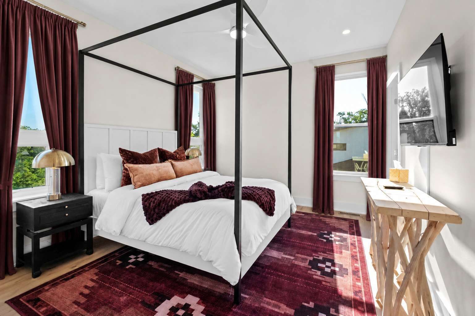 Una cama con dosel con un edredón blanco y almohadas marrones, una mesita de noche con una lámpara, un escritorio de madera con una silla y un ventanal con cortinas.