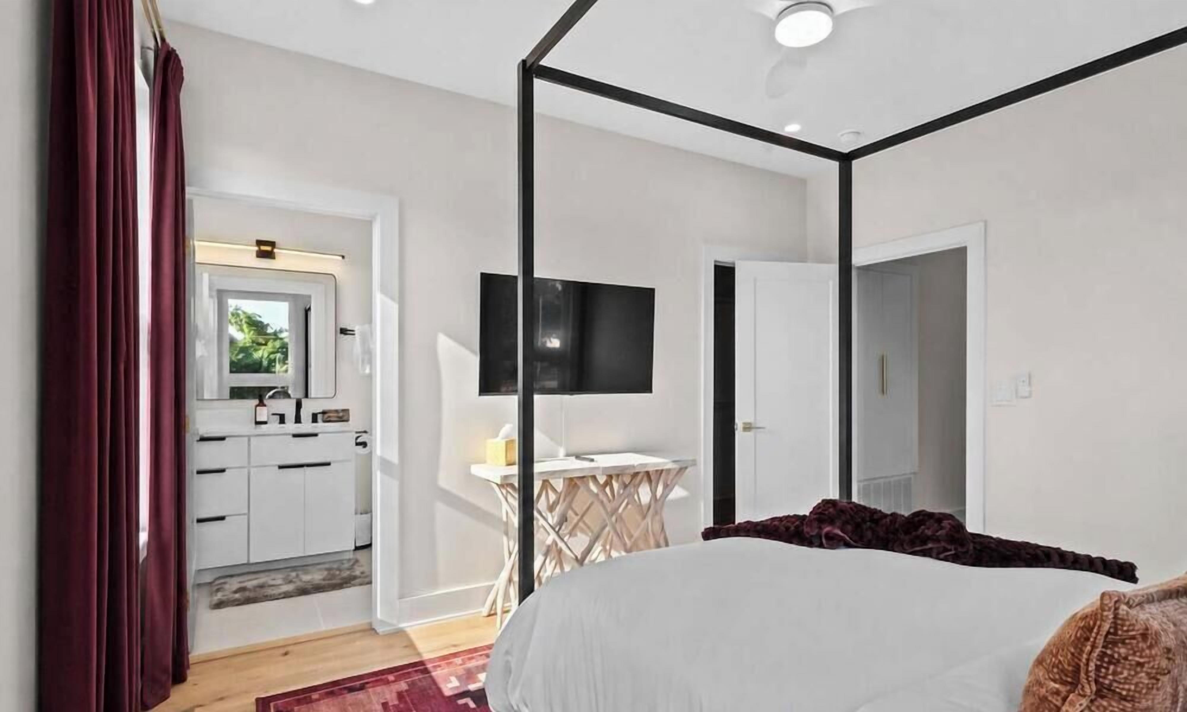 A bedroom with a four-poster bed, a desk with a flat-screen TV, and a view of a kitchen and bathroom through an open door.