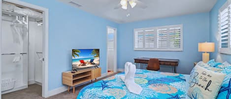 Ein Schlafzimmer mit Bett, Schreibtisch, Fernseher, Deckenventilator und einem Fenster mit Jalousien.