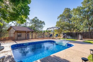 Una piscina rectangular amb un solàrium de fusta i una tanca, envoltada d'arbres i una casa.