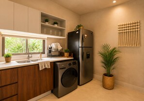A compact kitchen with a built-in washer and dryer, wooden cabinets, a window with a view, and a wall-mounted decorative item. - Modern Tulum Penthouse ● Private Plunge Pool ● Fast WiFi ● Gym (Tulum)