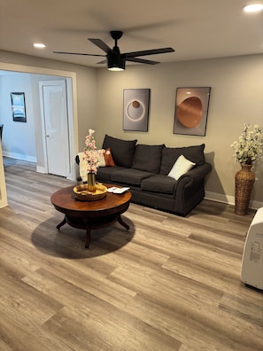 A living room with a dark grey sofa, a round wooden coffee table with a vase of flowers, a ceiling fan, and a door in the background.