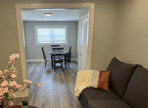 A living room with a dark gray sofa, a small table with a vase of pink flowers, and a dining area with a black table and chairs.