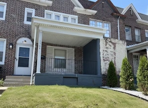 A two-story brick house with a covered porch, a front door, and a small front yard with grass and a few trees.