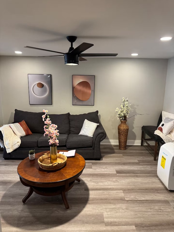 A modern living room with a dark grey sofa, a round wooden coffee table with a vase of flowers, and a ceiling fan.