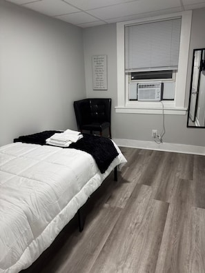 A bedroom with a bed, a chair, a window with blinds, and a wall-mounted air conditioner.