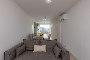 Un salon moderne avec un canapé gris, un coin repas comprenant une table et des chaises, et une vue sur l’extérieur.