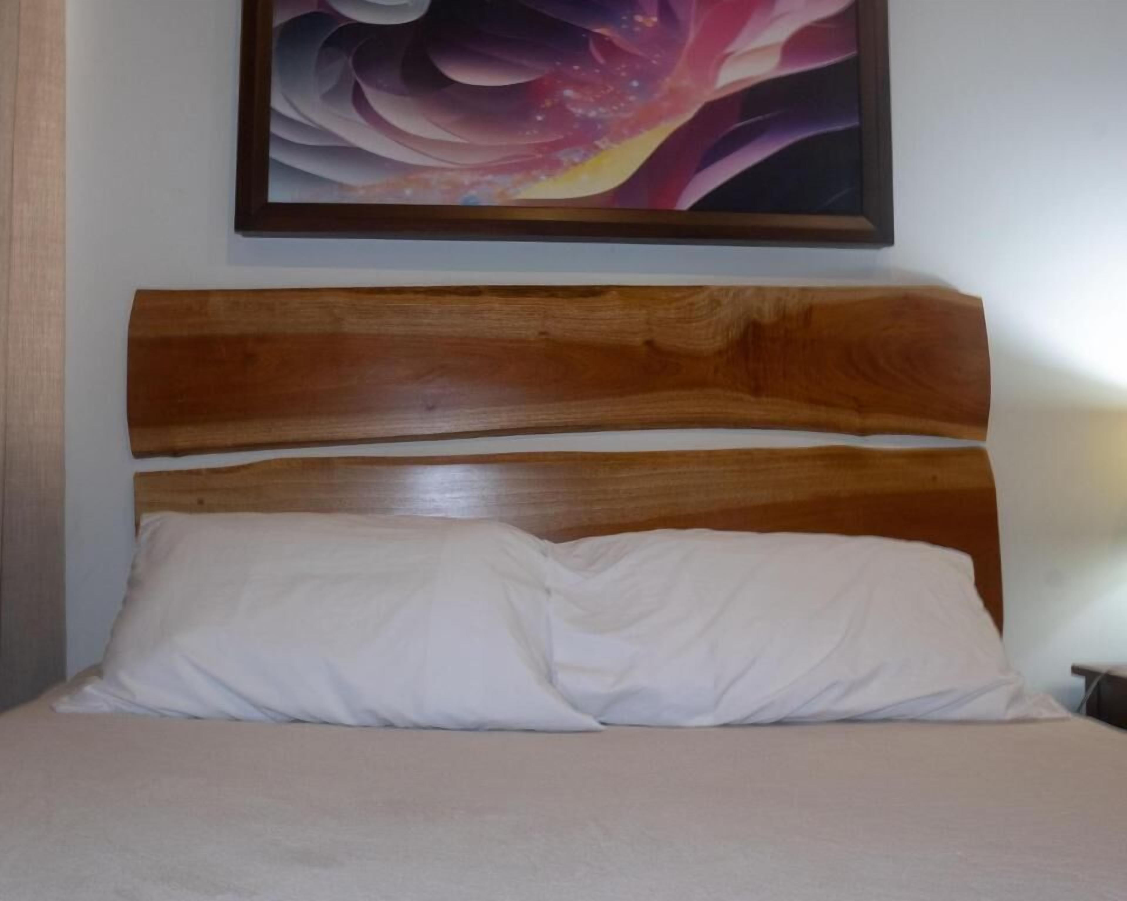 A bed with a wooden headboard, a white pillow, and a framed abstract painting above.