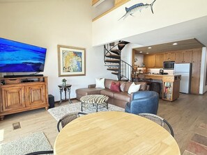 Uma sala ampla com televisão de tela plana, um sofá, uma mesa de centro e uma mesa de jantar. Uma escada em espiral leva a um andar superior com uma cozinha.