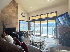 A living room with a fireplace, a large window with a view of the water, and a television on a stand. - Lake Chelan Shores Waterfront Dream #11-4 Pools Hot Tub Beach Tennis Boating (Chelan)