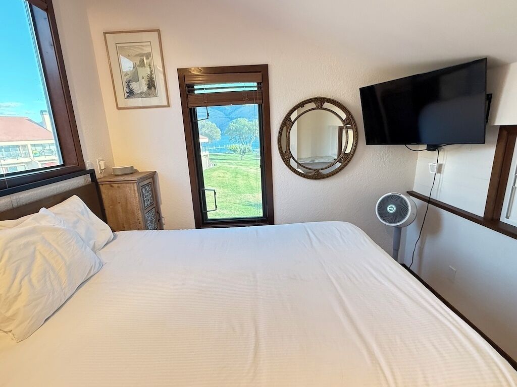 A hotel room with a bed, a television mounted on the wall, a window with a view of a green landscape, and a mirror on the wall.