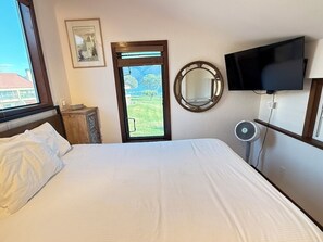 Quarto de hotel com uma cama, uma televisão fixada na parede, uma janela com vista para uma paisagem verde e um espelho na parede.