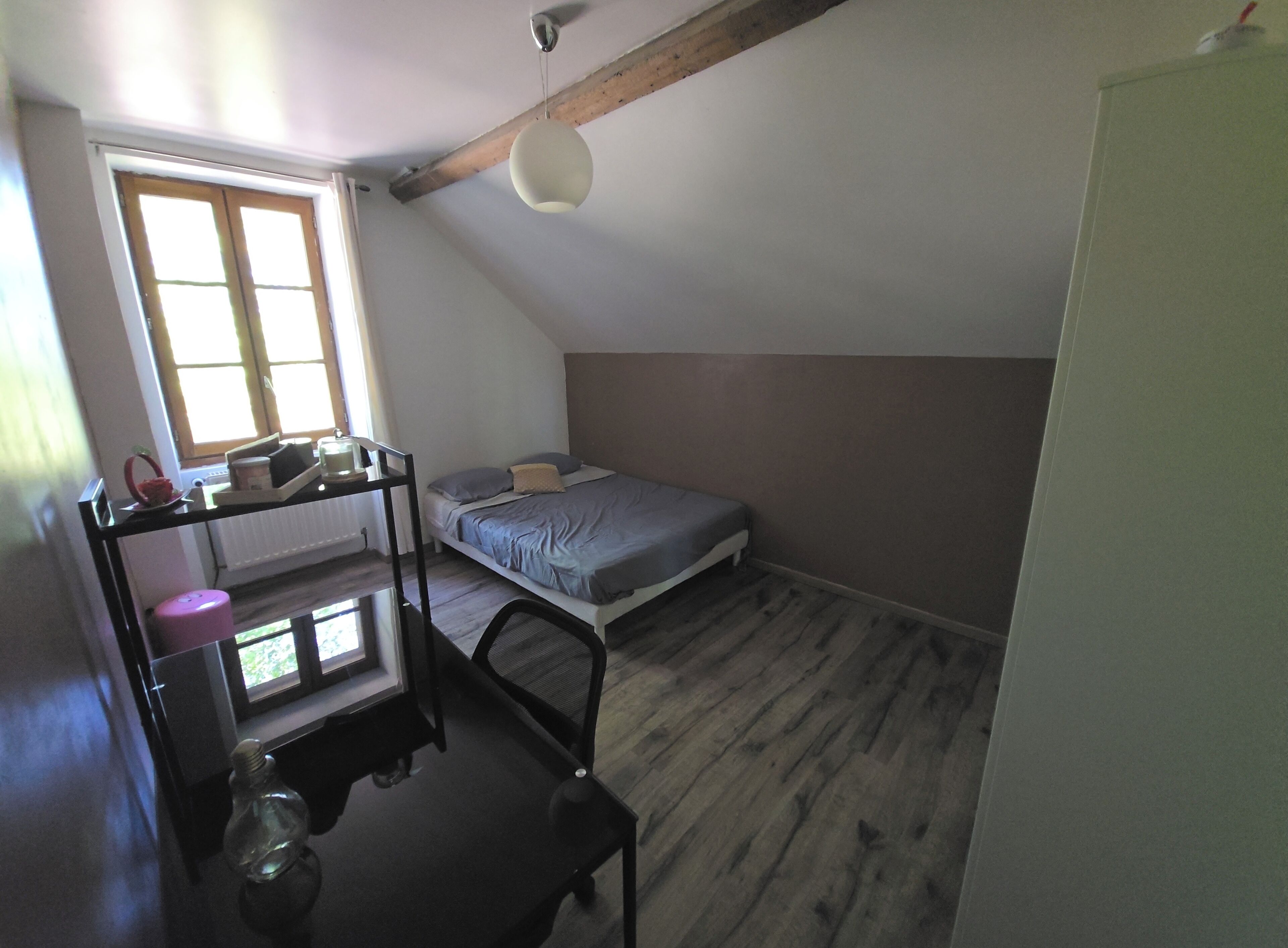 A small, single-room apartment with a sloped ceiling, a bed, a desk with a chair, and a window providing natural light.