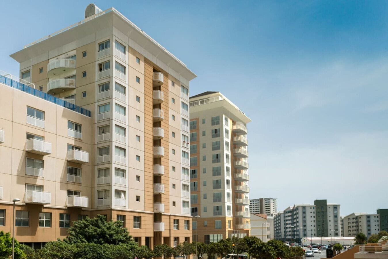 A multi-story residential building with balconies, a clear sky, and a street with vehicles and trees.