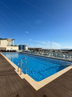 Ein Schwimmbecken mit einer Holzterrasse, im Hintergrund ein Yachthafen und ein klarer Himmel.