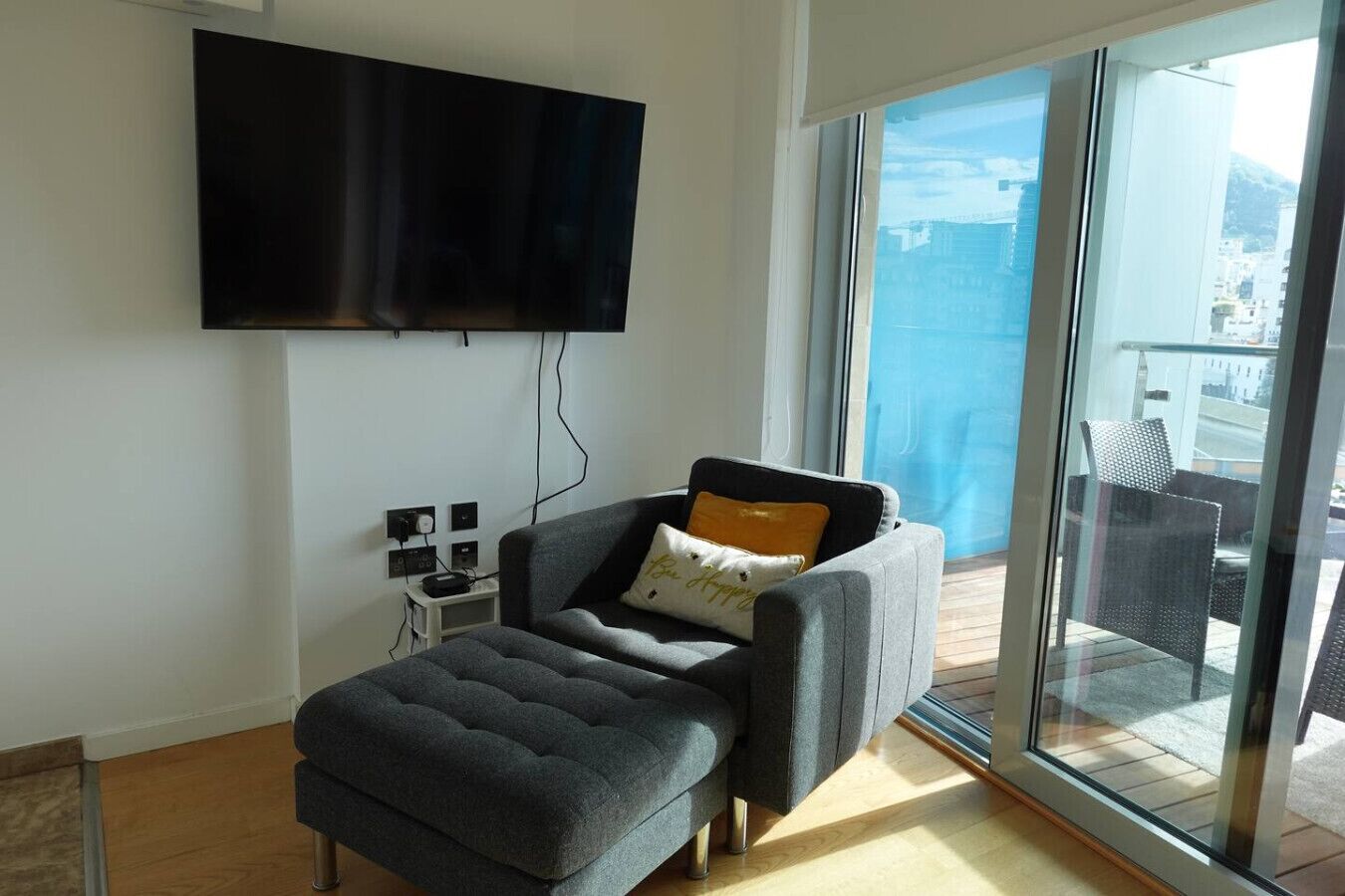 A modern living room with a flat-screen TV mounted on the wall, a grey sofa with a cushion, and a glass door leading to an outdoor balcony.