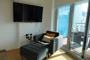 A modern living room with a flat-screen TV mounted on the wall, a grey sofa with a cushion, and a glass door leading to an outdoor balcony. - Gibraltar Luxury w/ Rooftop Pools & Views (Gibraltar)