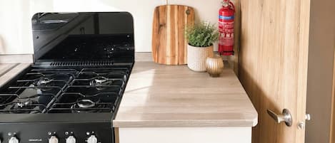A compact kitchen with a gas stove, wooden cabinets, and a built-in shelf with dishes and cups.