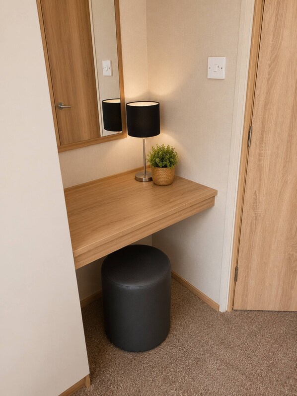 A room with a wooden desk, a black lamp, a small potted plant, a black ottoman, and a mirror on the wall.