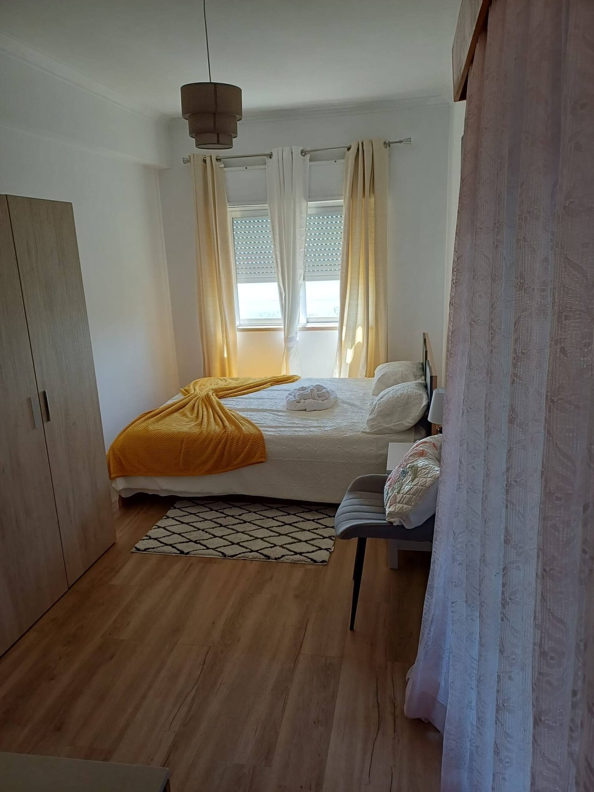 A bedroom with a bed, a wardrobe, a bedside table, a lamp, and a window with curtains.