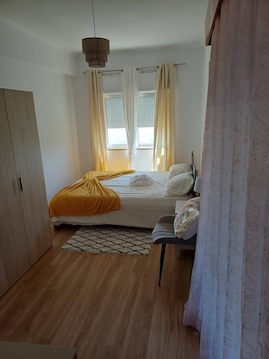 Um quarto com uma cama, guarda-roupa, mesa de cabeceira, abajur e uma janela com cortinas.
