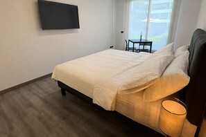 Ein Schlafzimmer mit einem Bett, einer Nachttischlampe, einem an der Wand montierten Fernseher und einem Fenster mit Blick ins Freie.