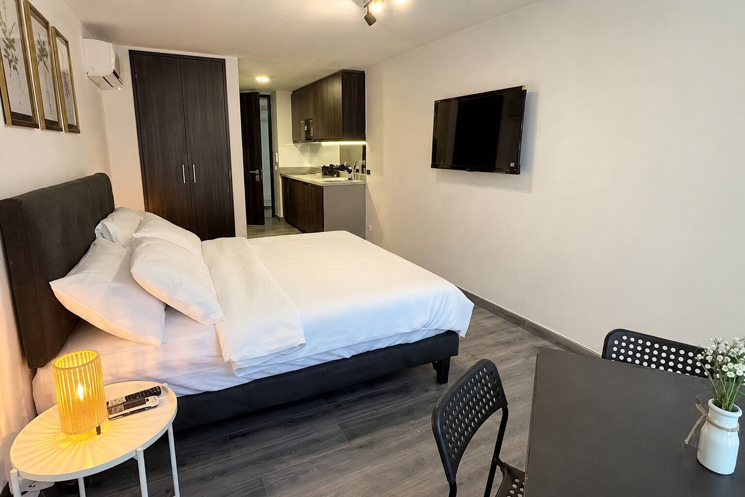 A hotel room with a bed, a small table with a lamp, a chair, a TV, and a kitchenette in the background.