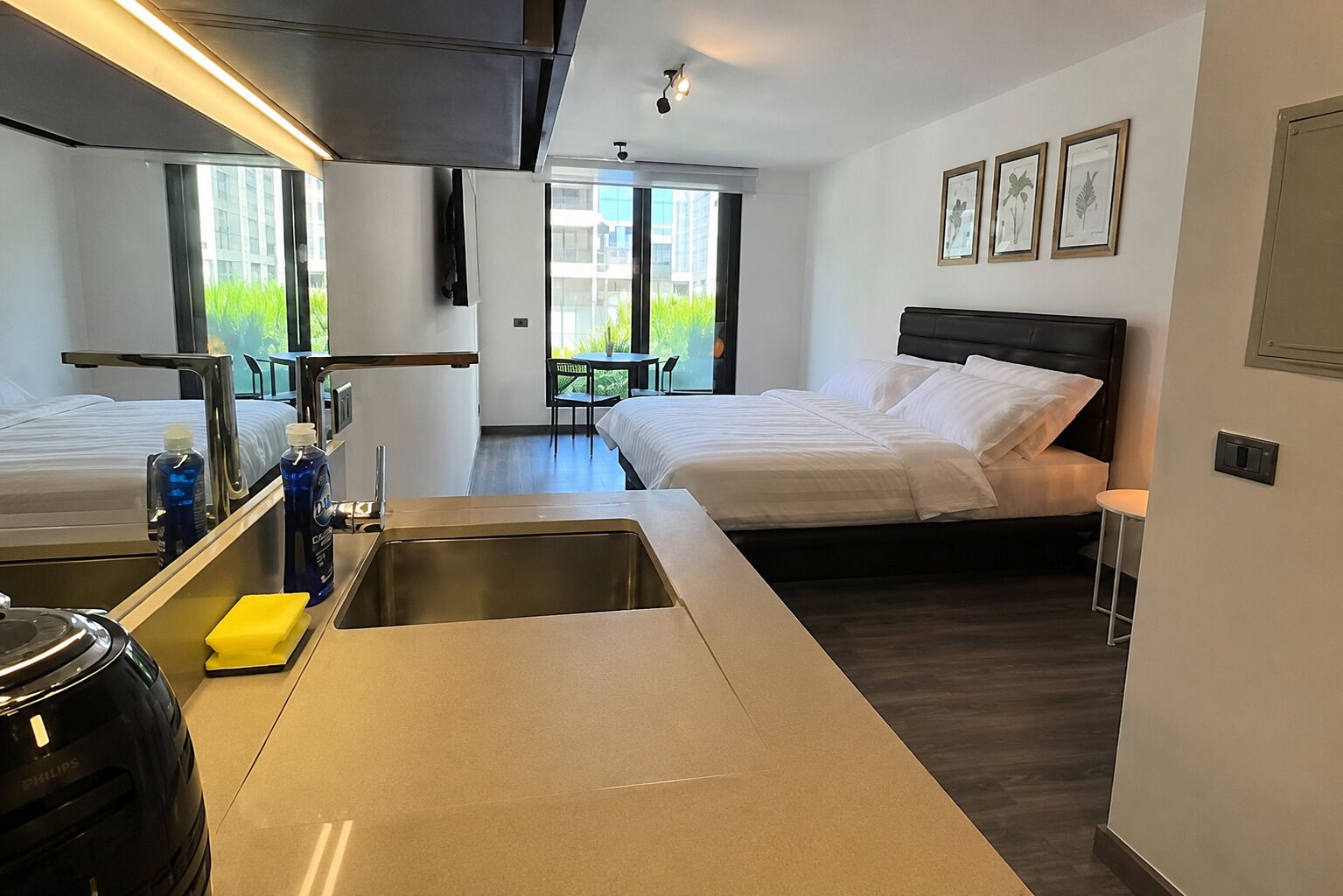 A modern hotel room with a kitchenette, a bed, a small table, and a view of greenery outside.