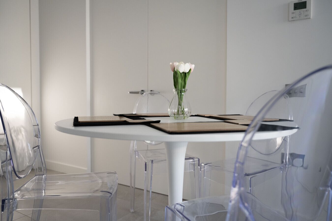 A room with a round table, clear chairs, a vase with flowers, and a thermostat on the wall.