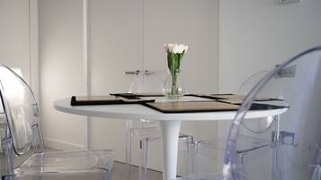 A room with a round table, clear chairs, a vase with flowers, and a thermostat on the wall.
