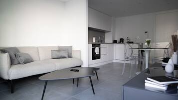 A modern living room with a sofa, coffee table, and clear chairs. A kitchen with white cabinets and appliances is visible in the background.