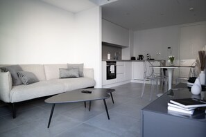 A modern living room with a sofa, coffee table, and clear chairs. A kitchen with white cabinets and appliances is visible in the background. - Skyline Studio EuroCity | Pool & Gym | City Centre (Gibraltar)