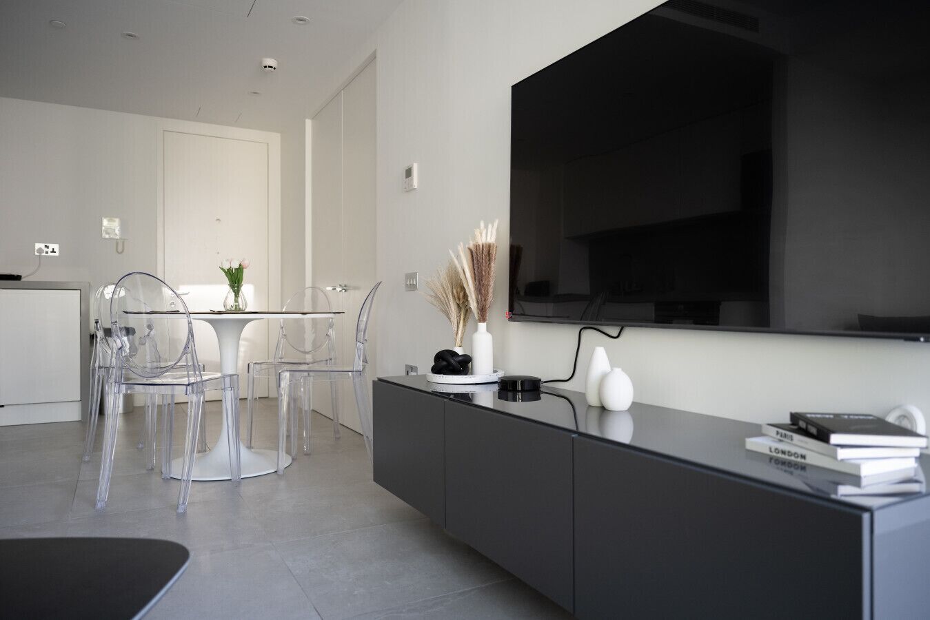 A modern living room with a large flat-screen TV mounted on the wall, a dining area with transparent chairs and a glass table, and a black console with books and decorative items.