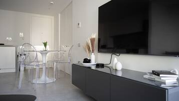 A modern living room with a large flat-screen TV mounted on the wall, a dining area with transparent chairs and a glass table, and a black console with books and decorative items.