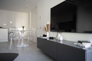 A modern living room with a large flat-screen TV mounted on the wall, a dining area with transparent chairs and a glass table, and a black console with books and decorative items. - Skyline Studio EuroCity | Pool & Gym | City Centre (Gibraltar)