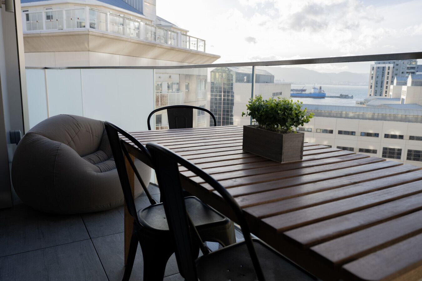 A balcony with a wooden table, two chairs, a bean bag, and a potted plant.