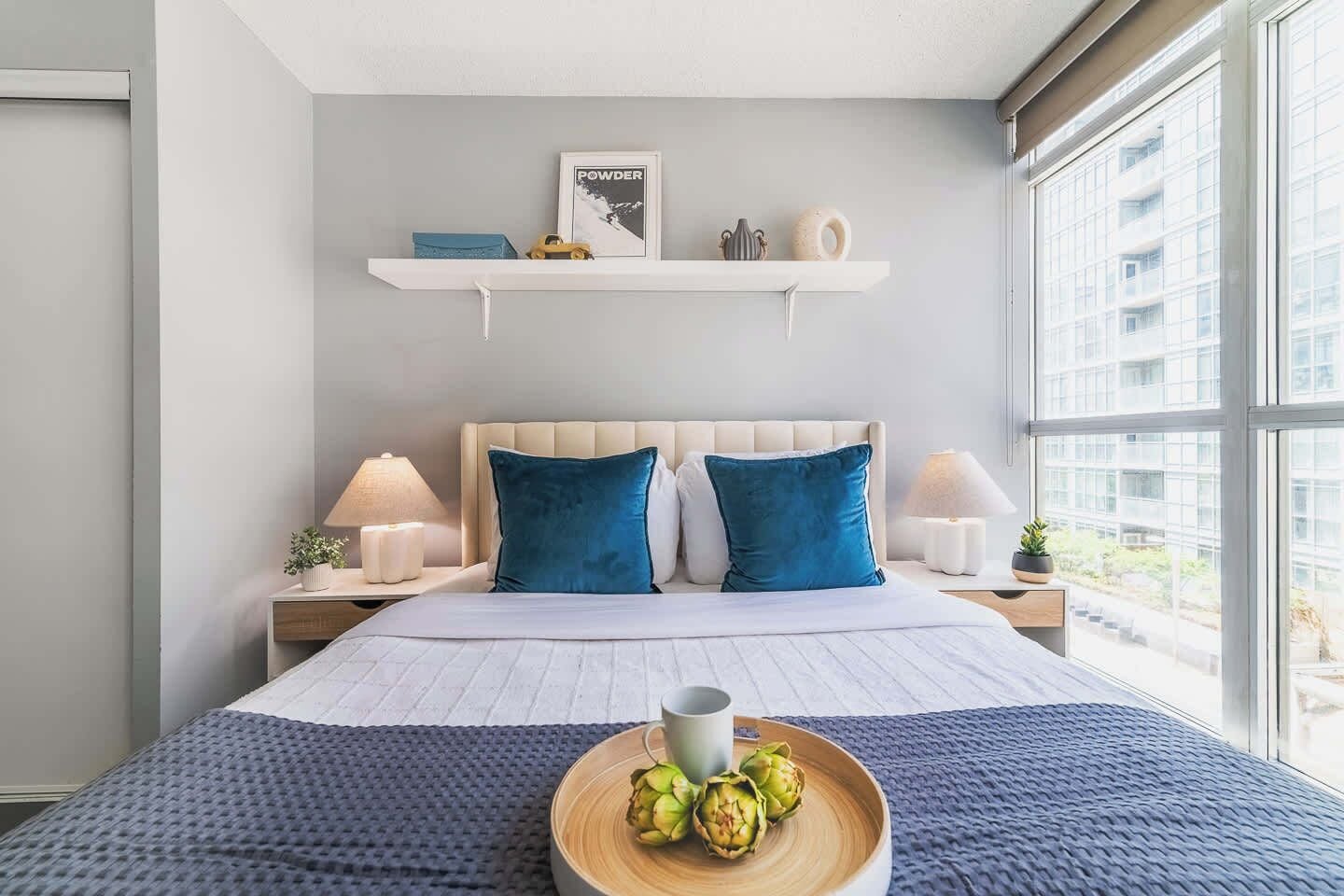A modern bedroom with a bed, bedside lamps, a window with a view, and a shelf with decorative items.